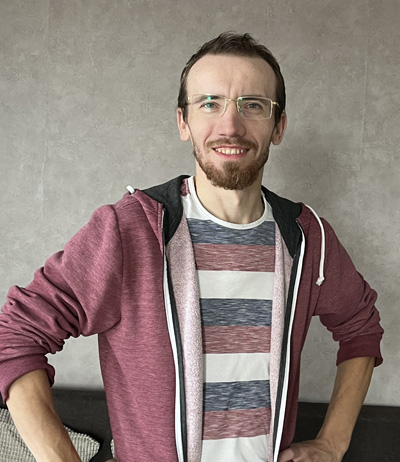 Uśmiechnięty mężczyzna w okularach, ubrany w bordową bluzę i koszulkę w paski, stoi z rękami opartymi na biodrach na tle jasnej ściany