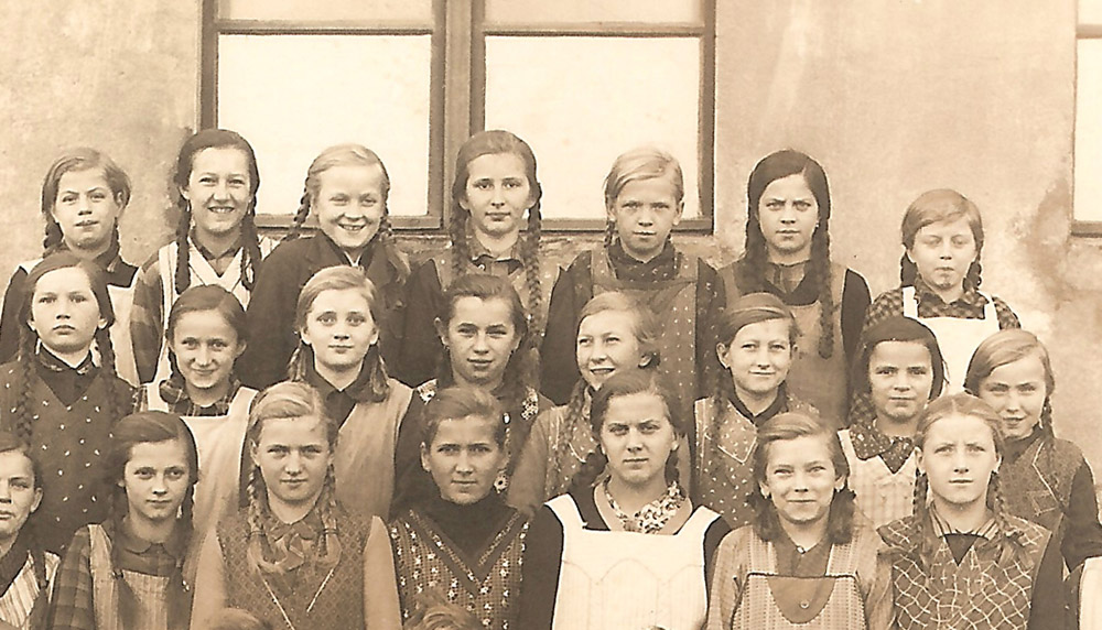 Grupowe zdjęcie szkolne dziewcząt stojących w kilku rzędach przed budynkiem z oknami, ubrane w stroje z epoki z warkoczami i poważnymi lub lekkimi uśmiechami na twarzach.